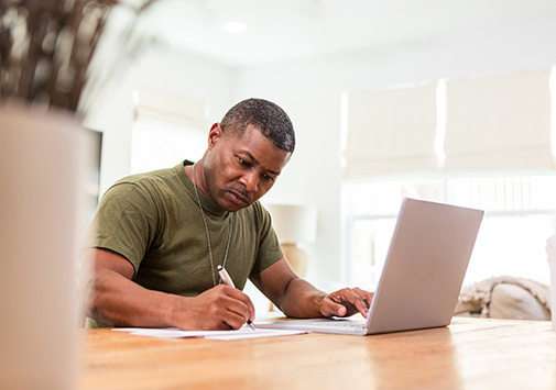 Military member sitting in his kitchen at his laptop, studying and writing notes.